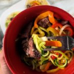 Red bowl full of zucchini noodles, sliced steak, and sliced bell peppers and onions.