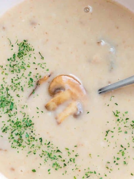 close up of a white bowl filled with cream of mushroom soup. THere is parsley sprinkled on the bowl of soup and a mushroom is visible under the broth.