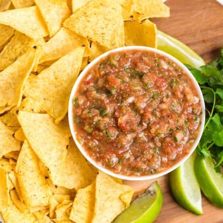 fresh salsa in a white bowl on a wood platter with tortilla chips, cilantro, and sliced limes in the background