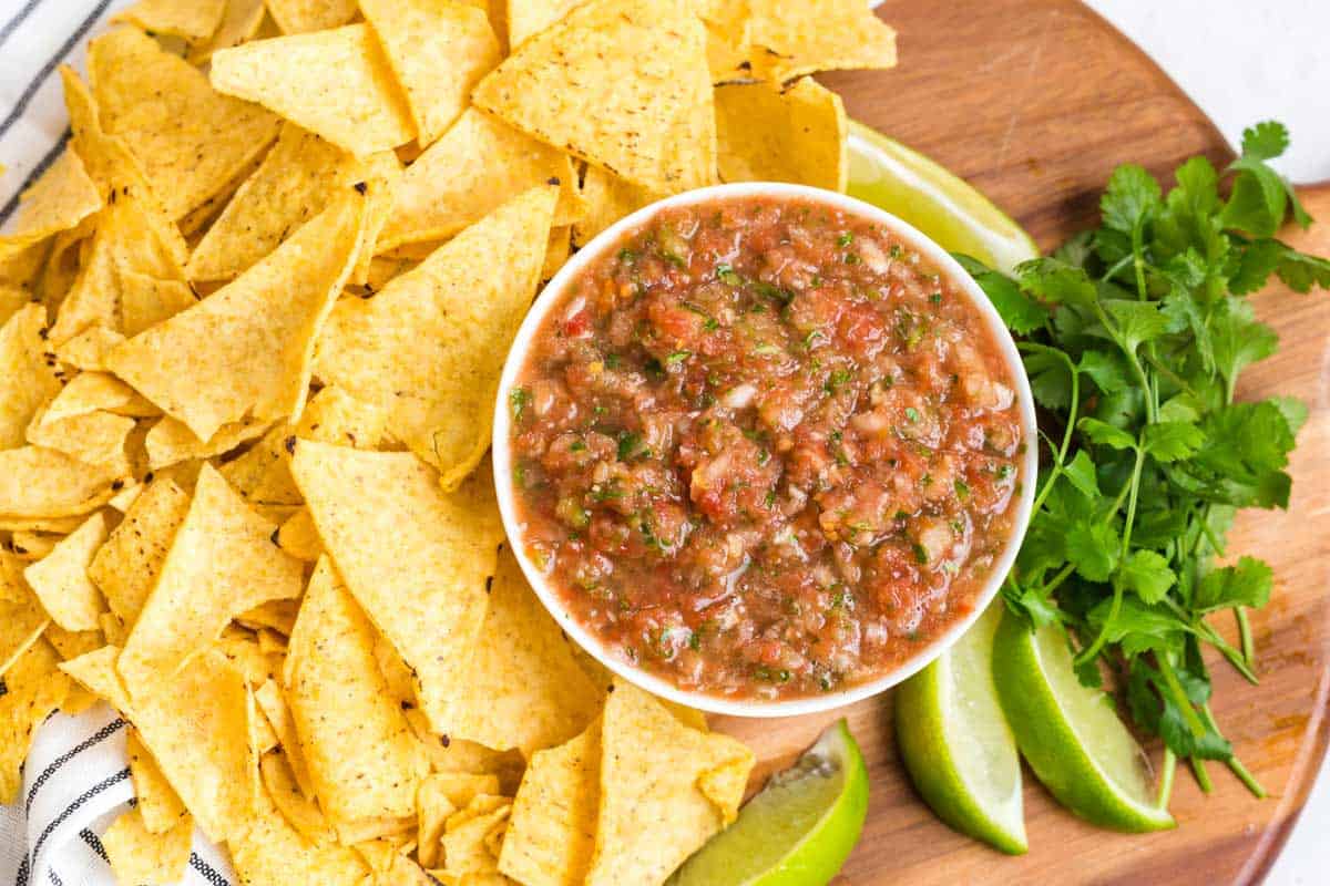 fresh salsa in a white bowl on a wood platter with tortilla chips, cilantro, and sliced limes in the background