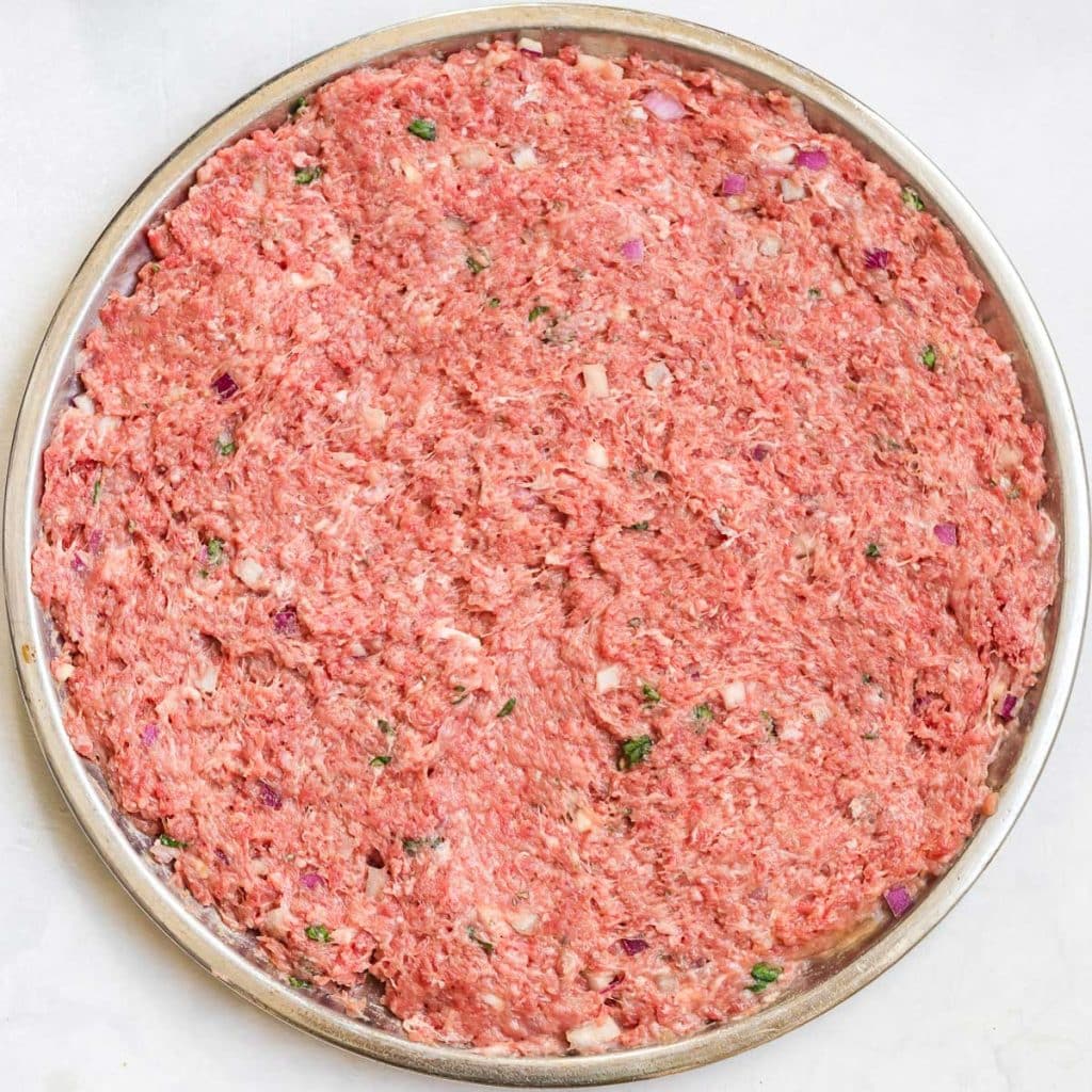 Silver pizza pan with uncooked ground meat and spices mixed in. 