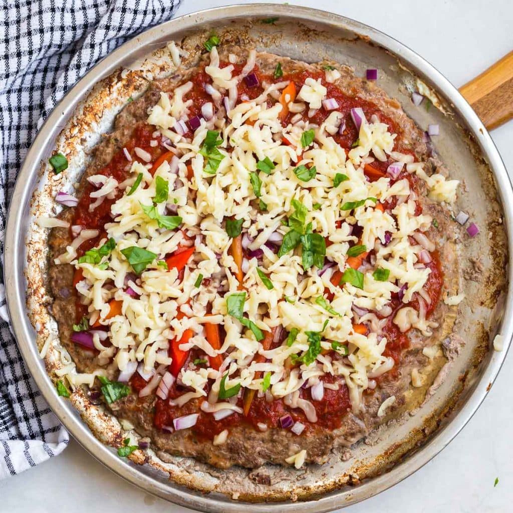 Silver pizza pan with cooked meat base and fresh toppings, getting ready to go into the oven.