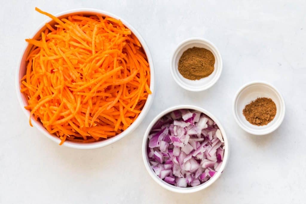 white bowls on a white counter filled with ingredients: shredded sweet potatoes, diced red onions, cinnamon and nutmeg. 