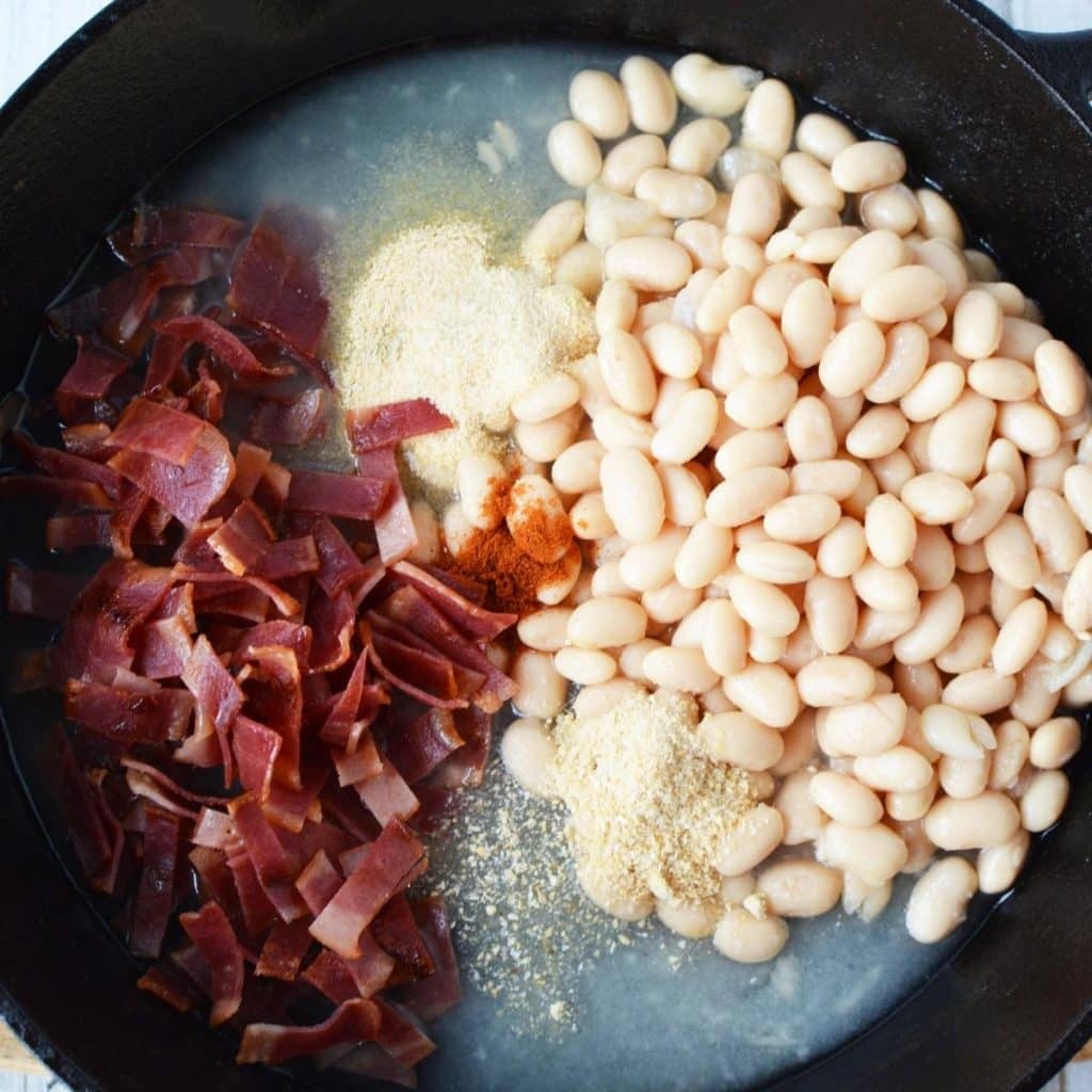 A black cast iron skillet with white beans, chopped and cooked bacon, and seasonings for Homemade Pork and Beans Recipe.