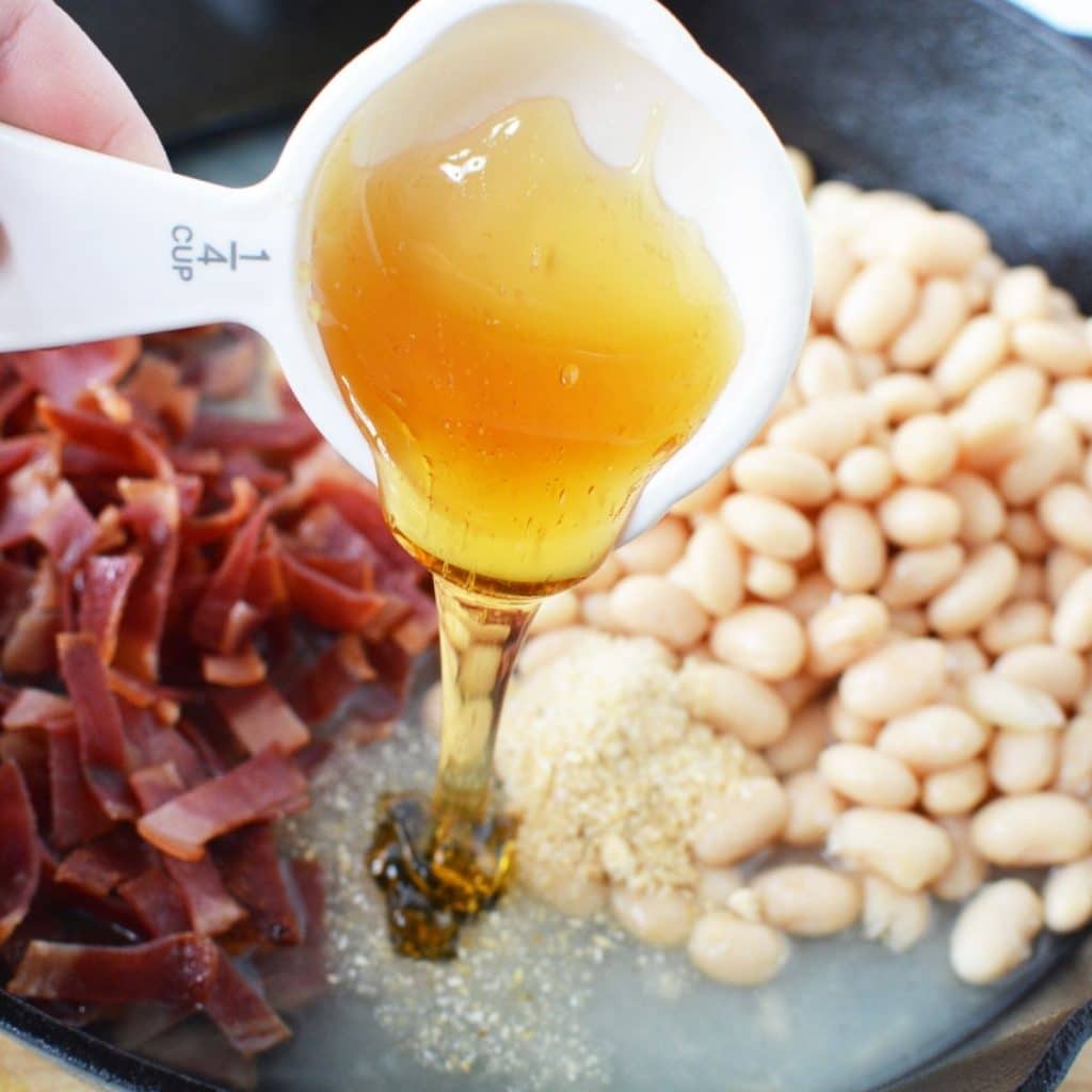 A cast iron skillet with chopped cooked bacon, white beans and a white measuring cup pouring honey into the skillet.