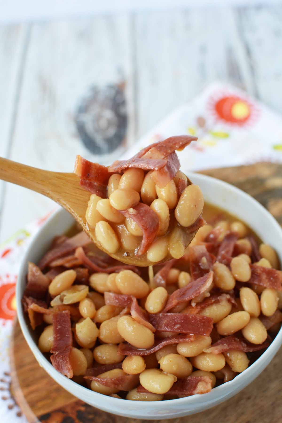 White bowl with cooked homemade pork and beans inside, there is a wooden spoon scooping some out. 