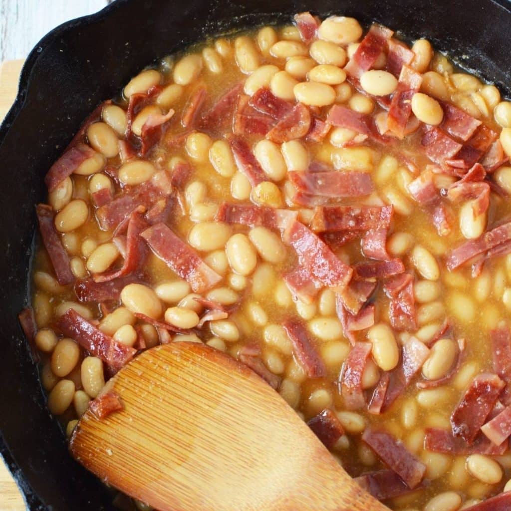 A black cast iron skillet with Homemade Pork and Beans being stirred by a wooden spoon.