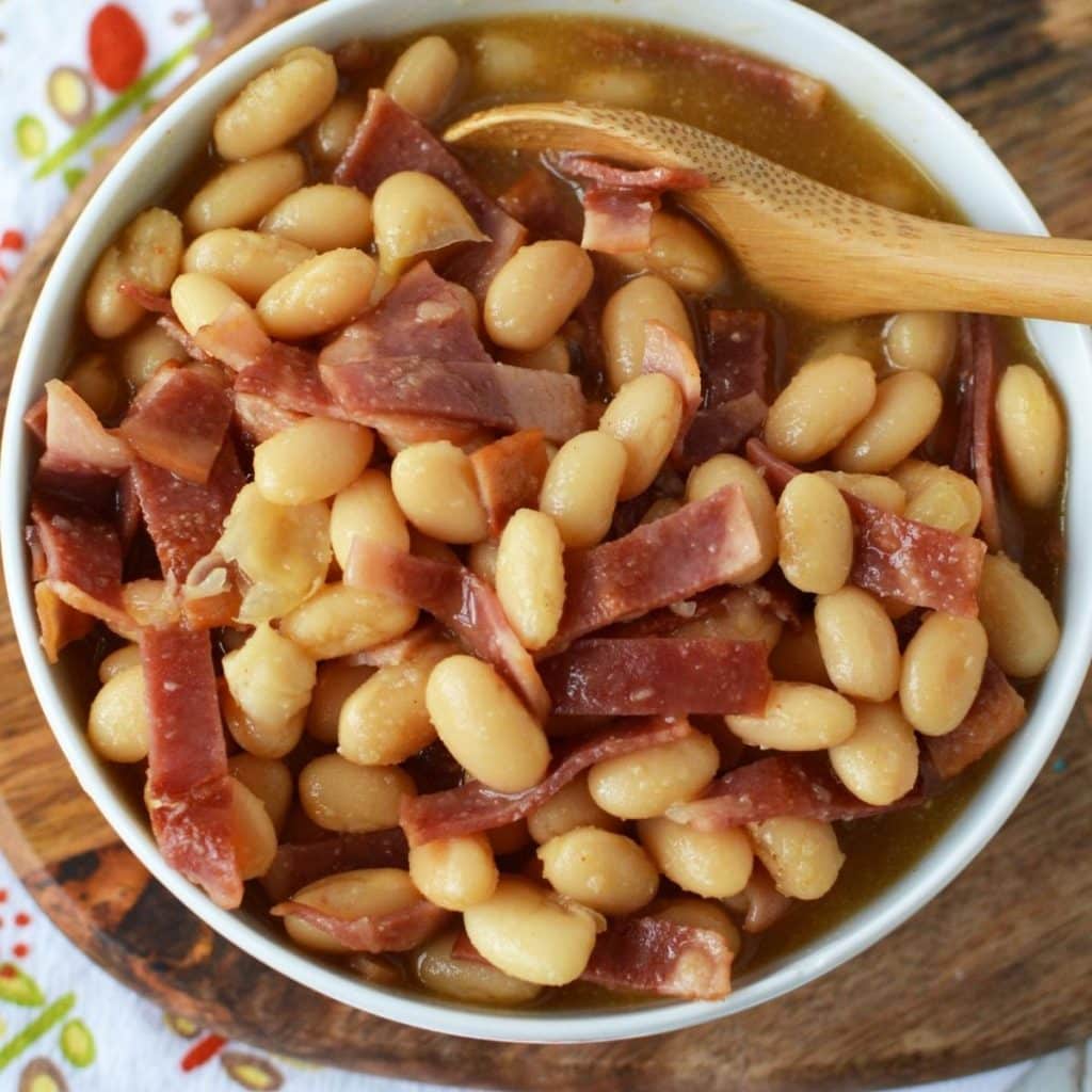 A white bowl with Homemade Pork and Beans being stirred by a wooden spoon.