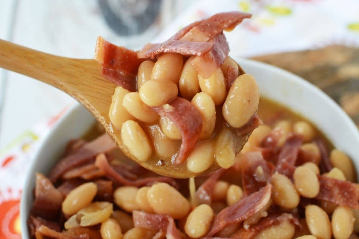 A white bowl filled with homemade pork and beans, with a wooden spoon lifting a hearty spoonful from the bowl.