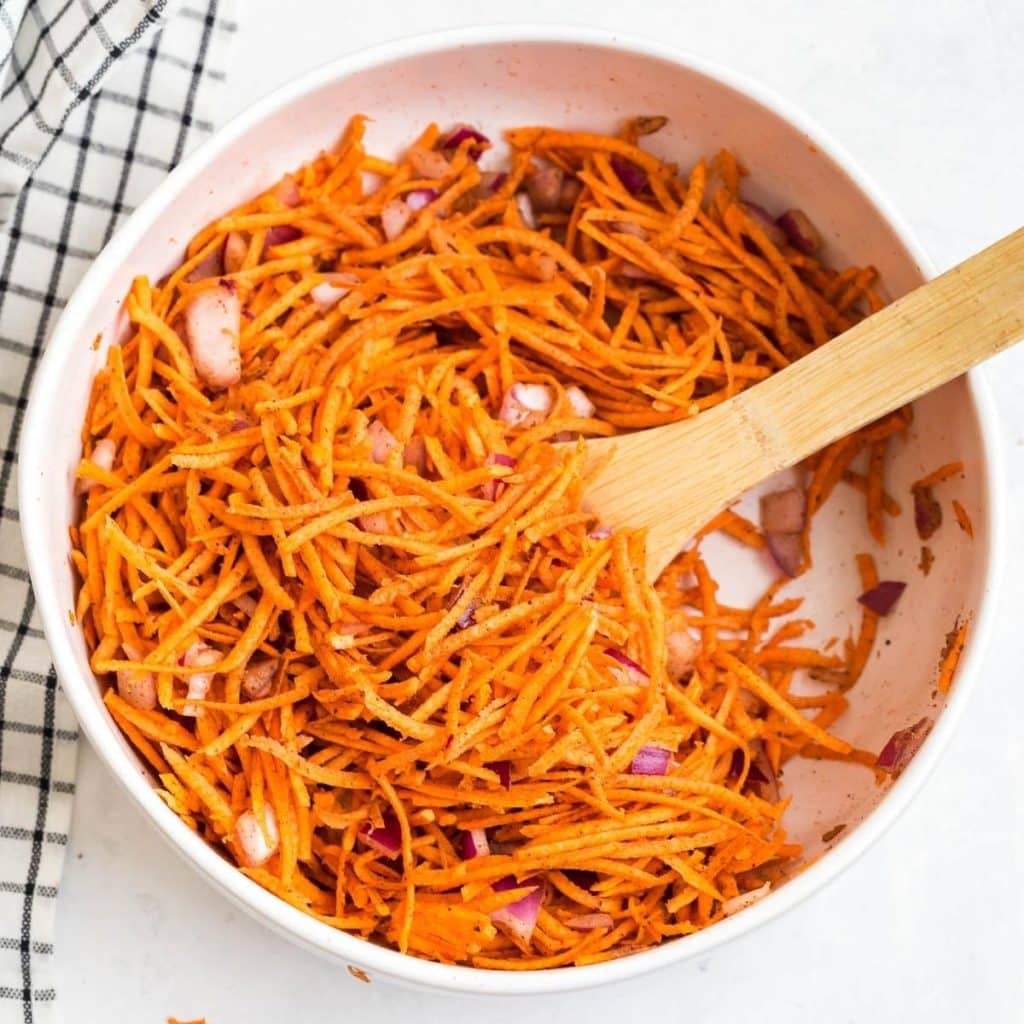 A white bowl with shredded sweet potatoes, diced red onion, cinnamon, and nutmeg all stirred together.