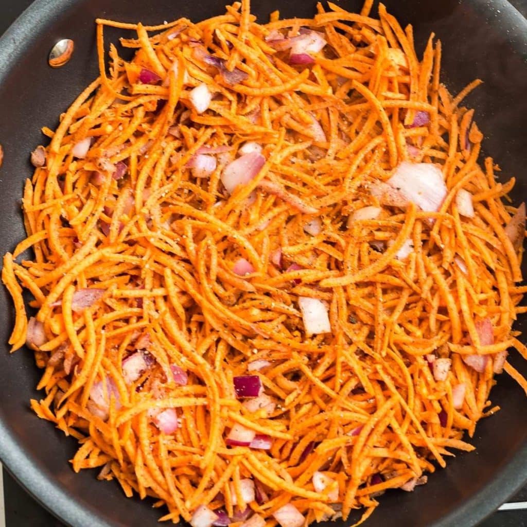 A black skillet with shredded sweet potatoes, diced onion and spices in the early stages of being cooked.
