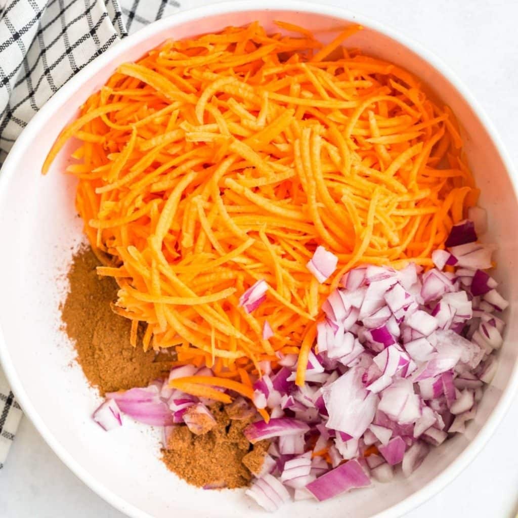 A white bowl with shredded sweet potatoes, diced red onion, cinnamon, and nutmeg before being stirred together.