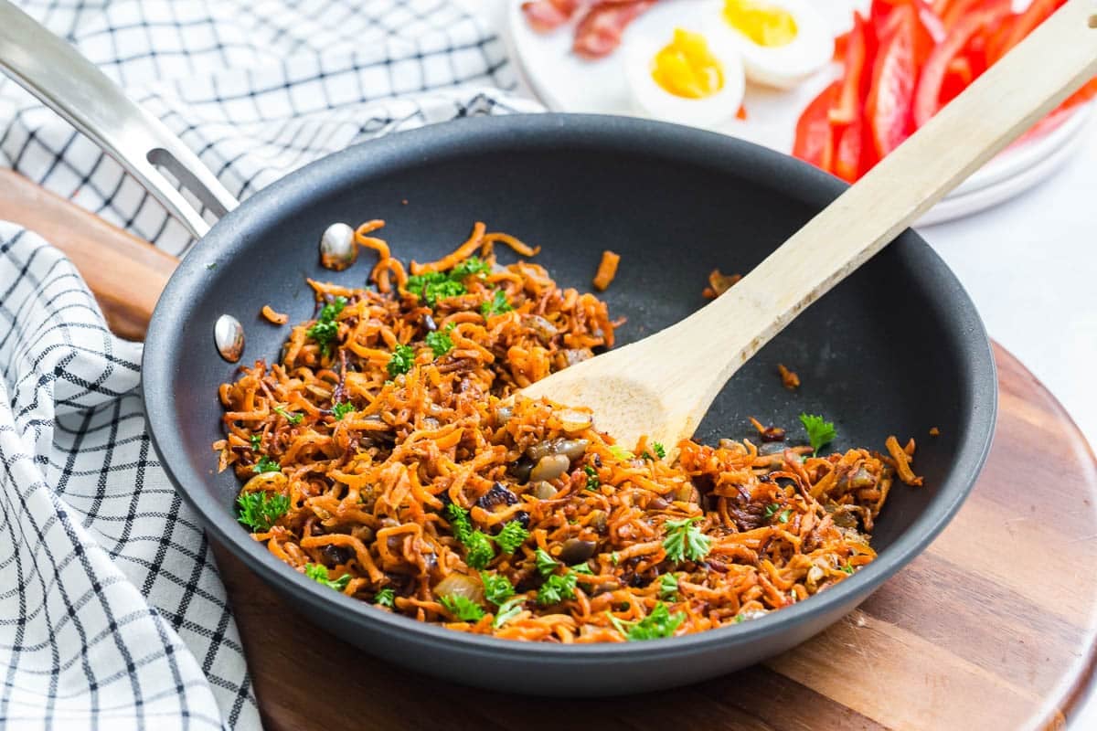Shredded sweet potatoes in a black skillet being mixed together by a wooden spoon.