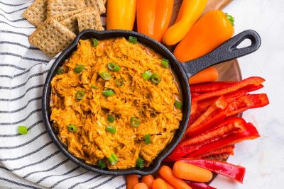 Buffalo chicken dip is in a small black cast iron skillet. Around the skillet are crackers, orange bell peppers, sliced red bell peppers, and baby carrots. The dip is garnished with sliced green onions.