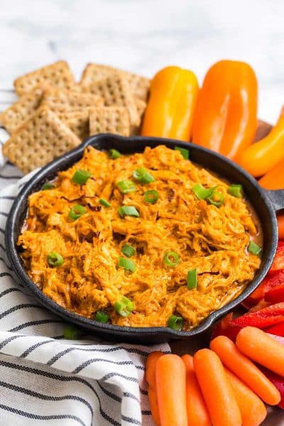 cast iron of buffalo chicken dip. Crackers, bell peppers, and carrots surround the cast iron skillet.