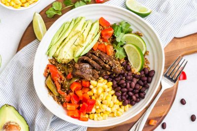 White bowl filled with beef fajitas and garnishes. Avocado, bell pepper, corn, black beans, and lime wedges.