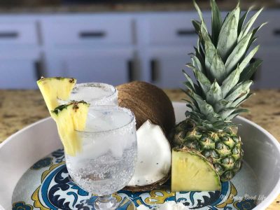 Pina colada margarita sits on a platter next to coconut parts and pineapple. The margarita is garnished with pineapple slices.
