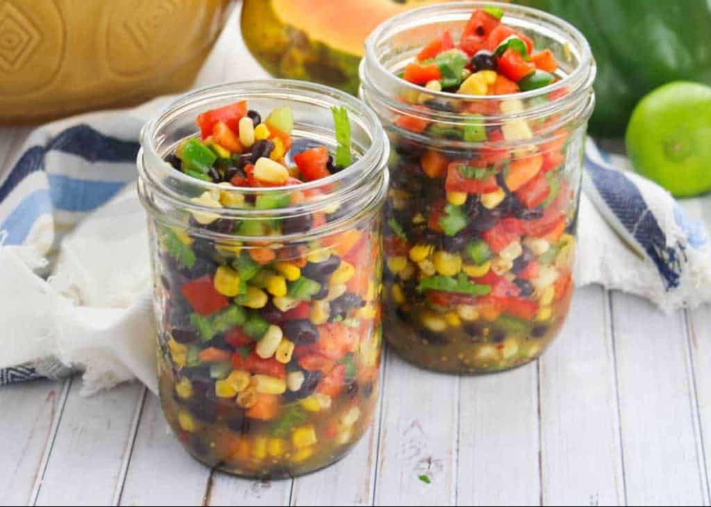 two small mason jars full of summer confetti salad with fresh food in the background