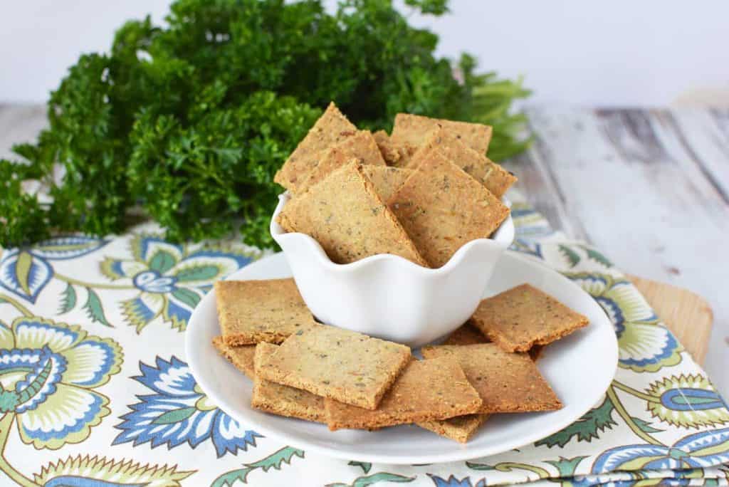Image of a white bowl with healthy crackers. Under the bowl is a white plate with crackers arranged around the bowl. In the background there is a bunch of parsley. 