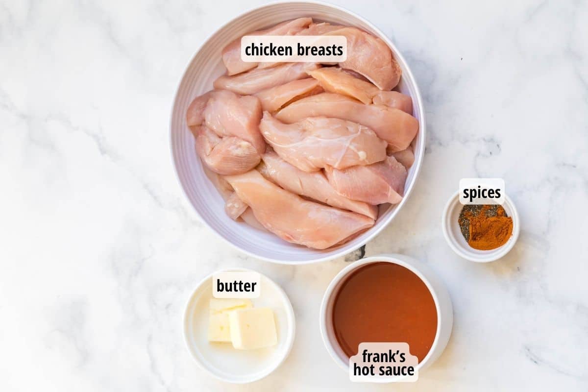 A white and gray marbled counter with labeled bowls full of chicken breasts, butter, hot sauce and spices.