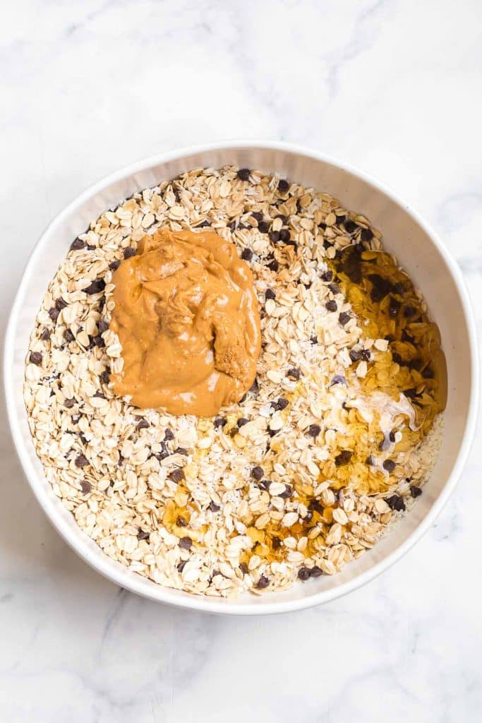 white bowl with mixed together oats, chocolate chips, coconut, and topped with peanut butter and honey to be mixed in