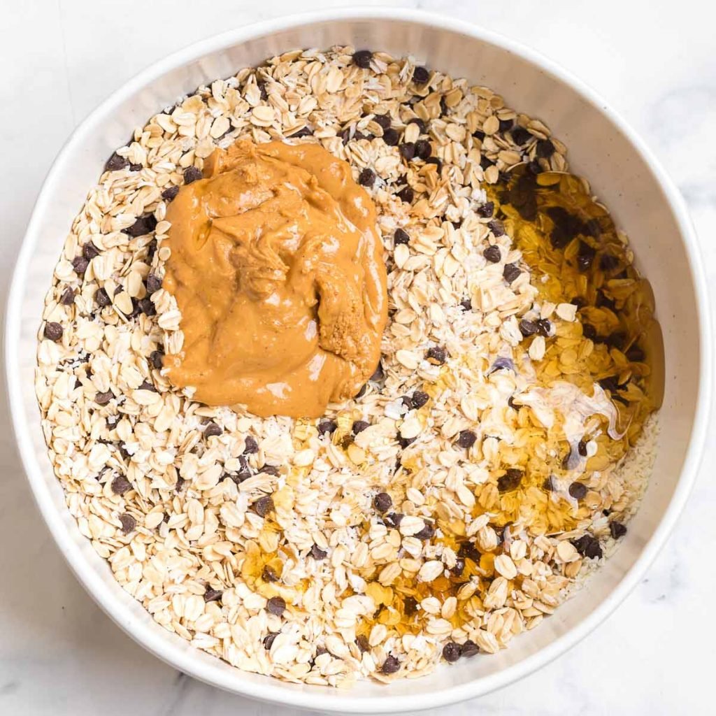 white bowl with mixed together oats, chocolate chips, coconut, and topped with peanut butter and honey to be mixed in