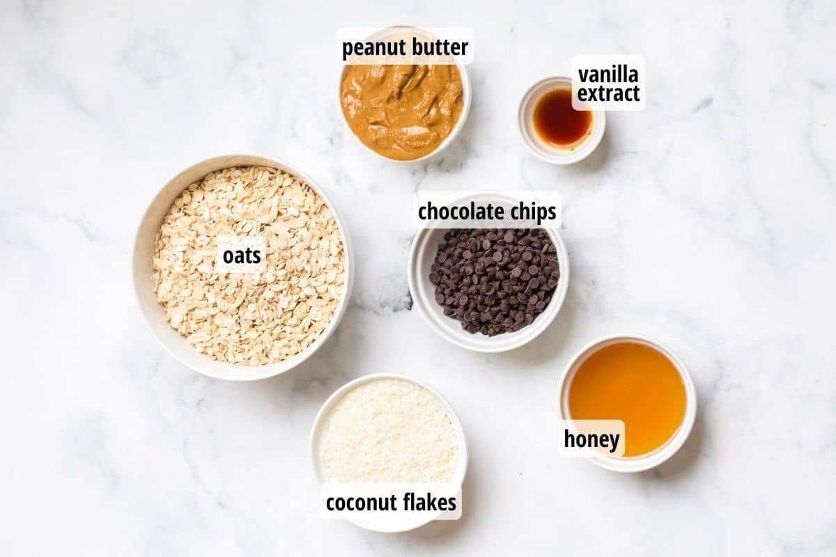 A marble counter top with bowls full of labeled ingredients including oats, coconut flakes, honey, chocolate chips, vanilla extract and peanut butter.