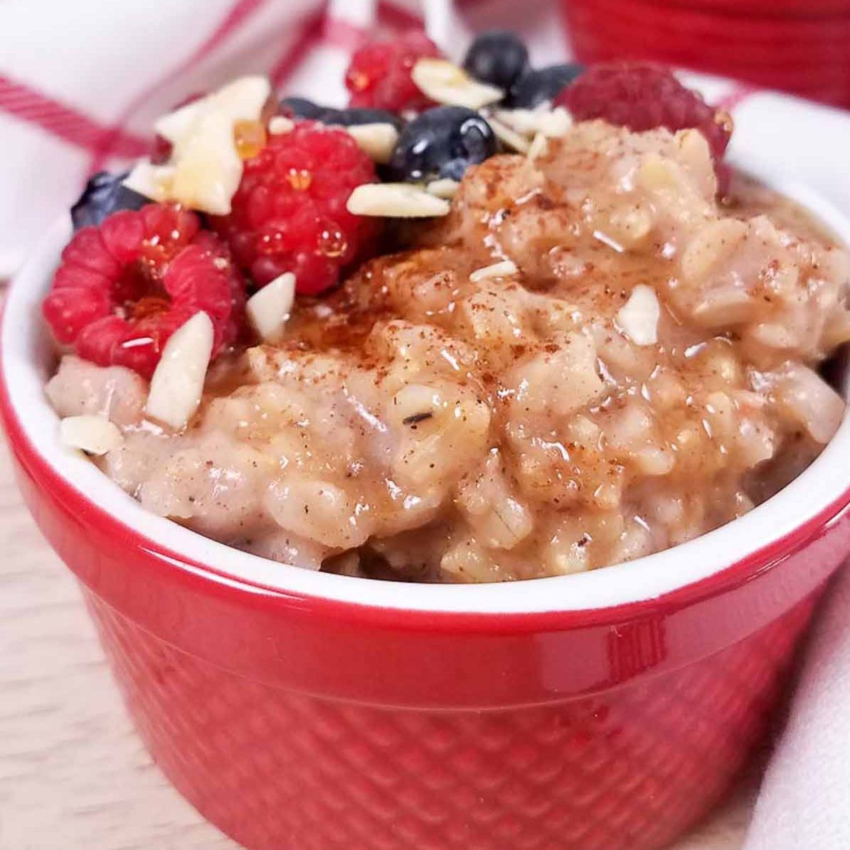 Red ramekin with cooked rice pudding filling it. There are raspberries, blueberries, and slivers of almonds on the left side of the bowl. 