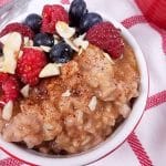 A red ramekin full of healthy rice pudding. There are berries and slivered almonds on top of the ramekin and a white and red striped napkin underneath.