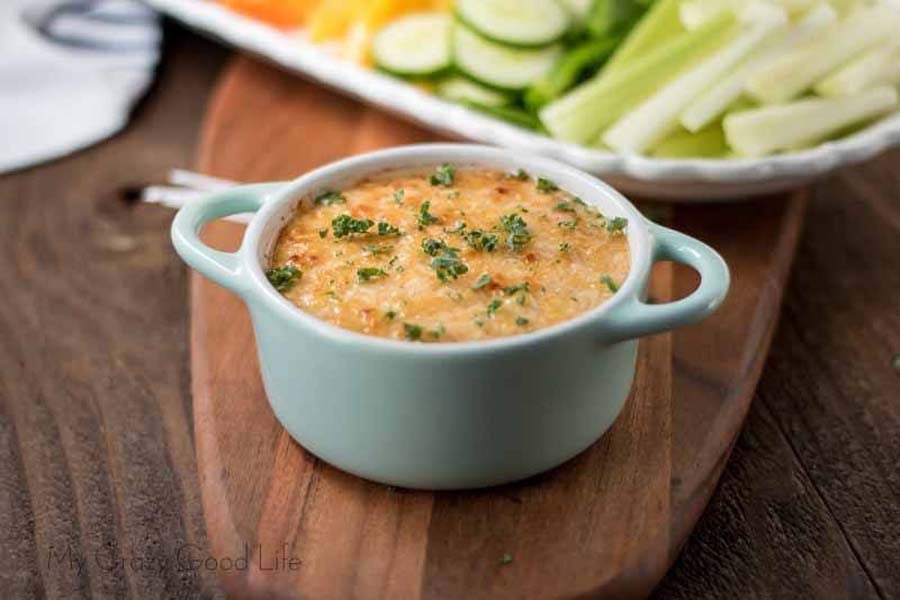 Baked Buffalo Chicken Dip in a white ramekin with two handles in front of a tray of fresh cut mixed veggies.
