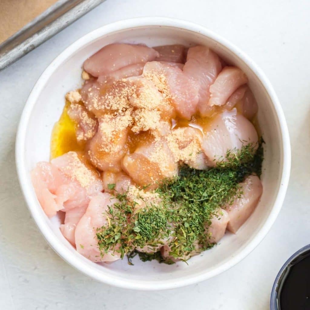 A white bowl with diced chicken, oil and seasonings in it.