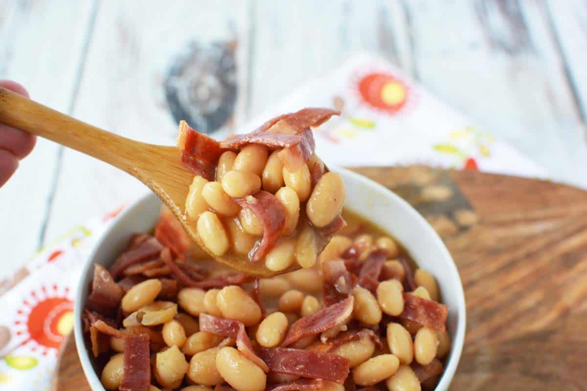 White bowl of healthy pork and beans with a small wooden ladle holding up a spoonful.