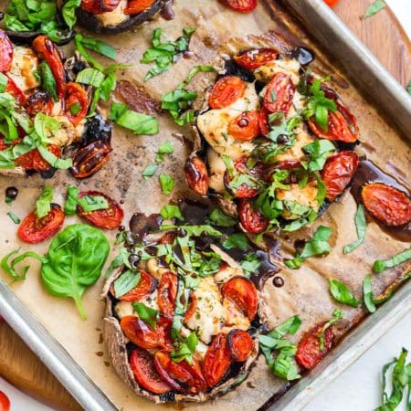 Four portobello mushrooms on a baking sheet that have been cooked with chicken and tomatoes and topped with fresh basil and a balsamic reduction.