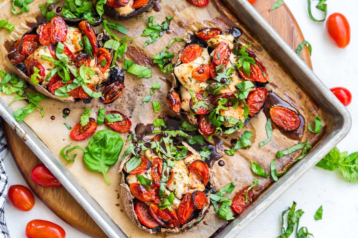 Four portobello mushrooms on a baking sheet that have been cooked with chicken and tomatoes and topped with fresh basil and a balsamic reduction.