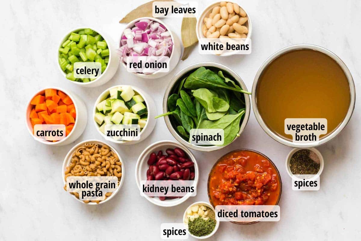 Ingredients for Minestrone soup in white and clear bowls laid out on a white countertop. 