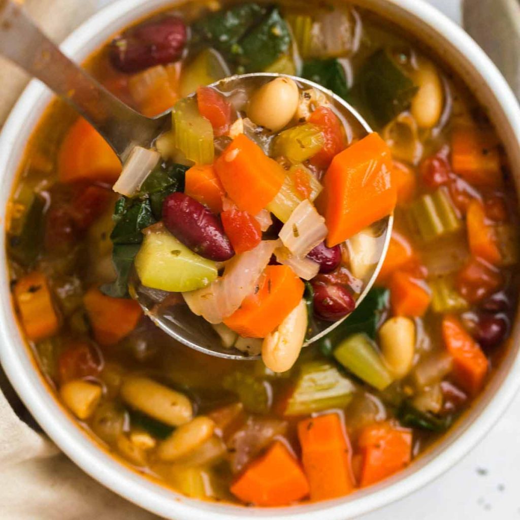 White bowl filled with Minestrone soup. There is a spoon lifting soup up towards the camera.
