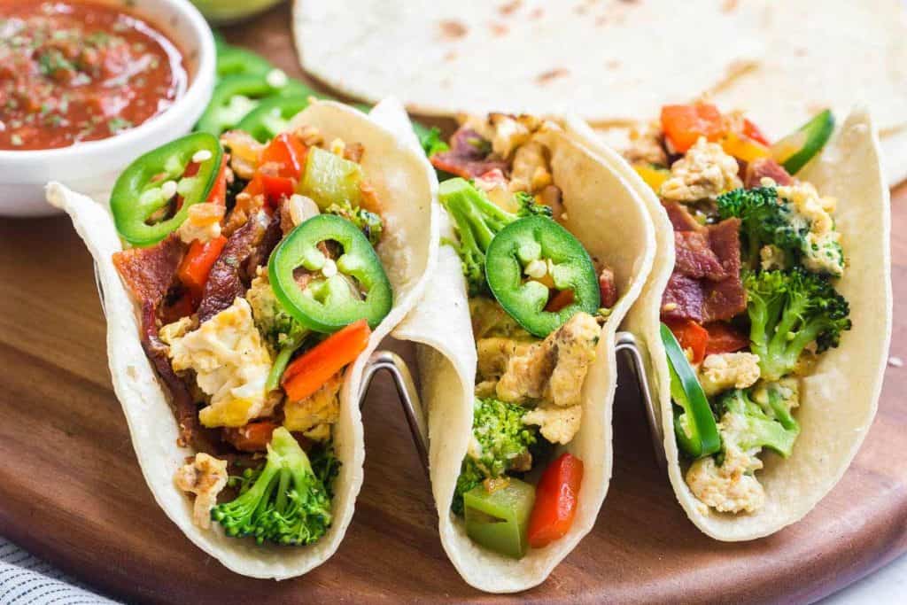 three breakfast tacos with egg, bacon, broccoli and jalapeños on a wood platter.