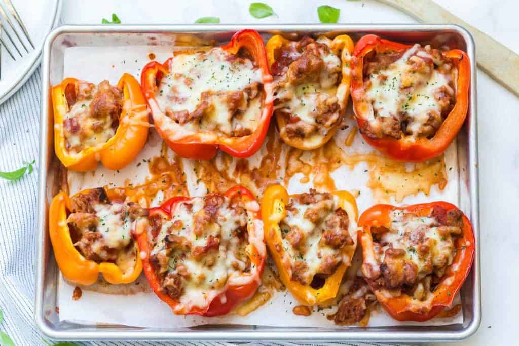 Image of red and orange pizza stuffed bell peppers on a metal baking sheet. Melted cheese is sprinkled on top of the peppers.