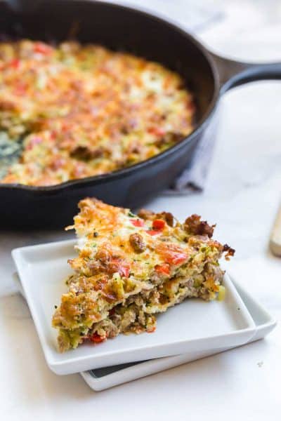 Slice of zucchini sausage breakfast casserole sits on square white plate. In the background there is a cast iron skillet with the remaining portion of the breakfast casserole.