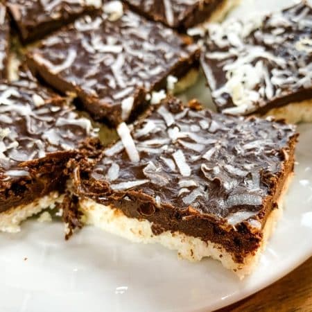 A white plate with cut coconut mocha bars on it. The bars have a cream colored coconut base, chocolate middle and are topped with coconut flakes.