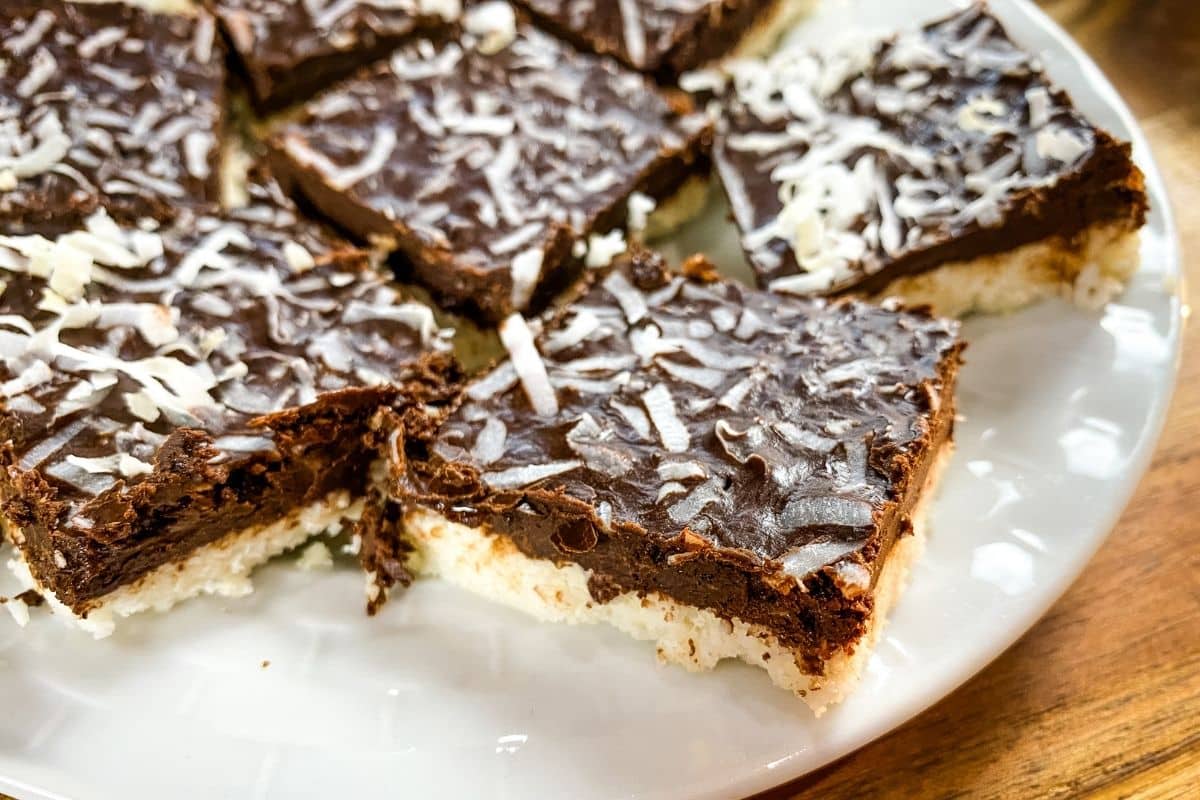 A white plate with cut coconut mocha bars on it. The bars have a cream colored coconut base, chocolate middle and are topped with coconut flakes. 