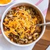 A white bowl filled with goulash, topped with shredded cheese, and sitting on top of a red and white napkin that's placed on a circular wooded board.