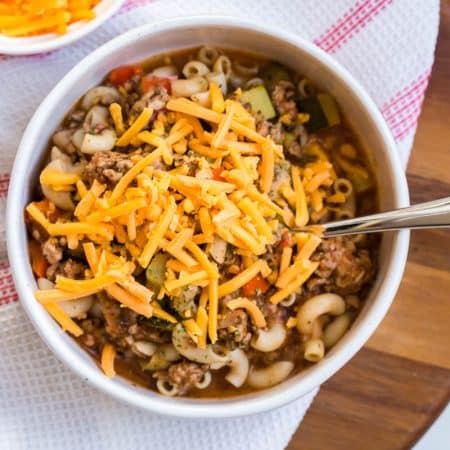 A white bowl filled with goulash, topped with shredded cheese, and sitting on top of a red and white napkin that's placed on a circular wooded board.
