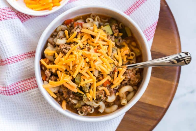 A white bowl filled with goulash, topped with shredded cheese, and sitting on top of a red and white napkin that's placed on a circular wooded board.