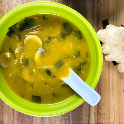 Miso soup in a green bowl with fresh ginger and garlic off to the side.