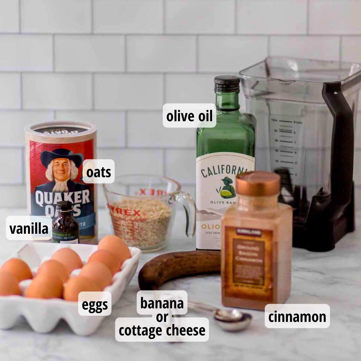 Ingredients for pancakes without flour on a white counter: a carton of eggs, container of oats, vanilla, cinnamon, banana, and olive oil.