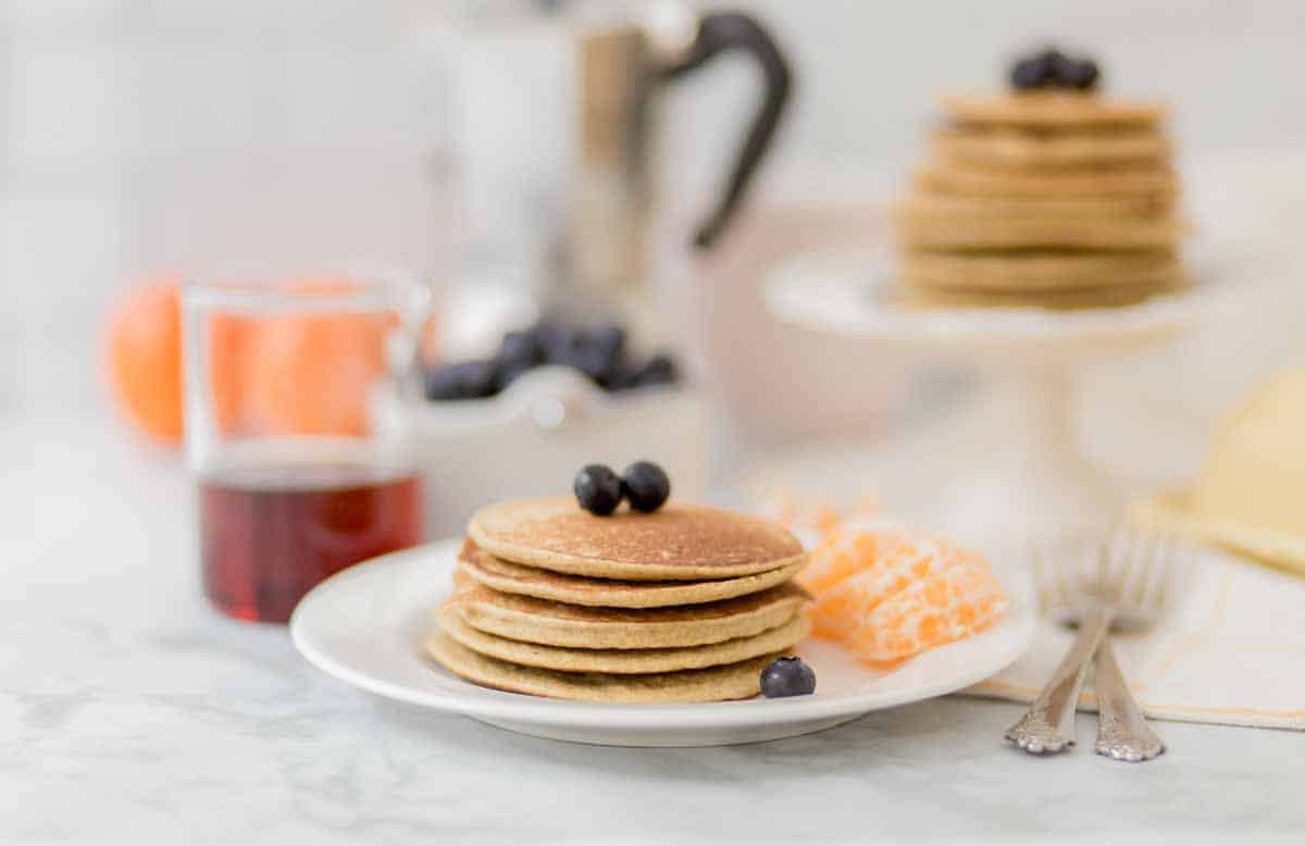 Stack of cooked pancakes on top of a white plate. There are blueberries and orange slices on the plate as well. 