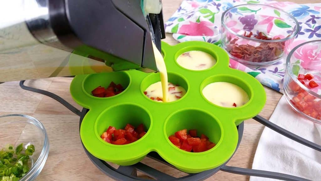 blender pouring egg mixture into a green egg bite mold filled with red peppers and green onions
