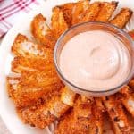 A white bowl with a cut and air fried blooming onion and in the center is a glass bowl of spicy dipping sauce.