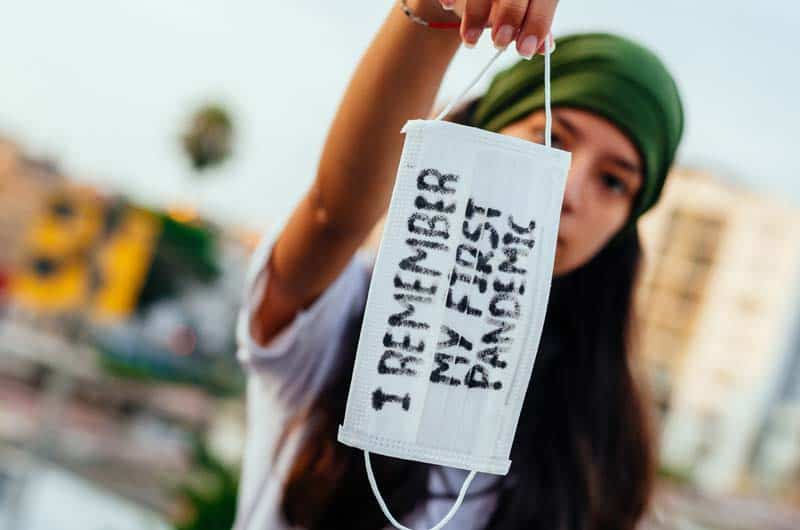 girl holding up face mask that reads "i remember my first pandemic"